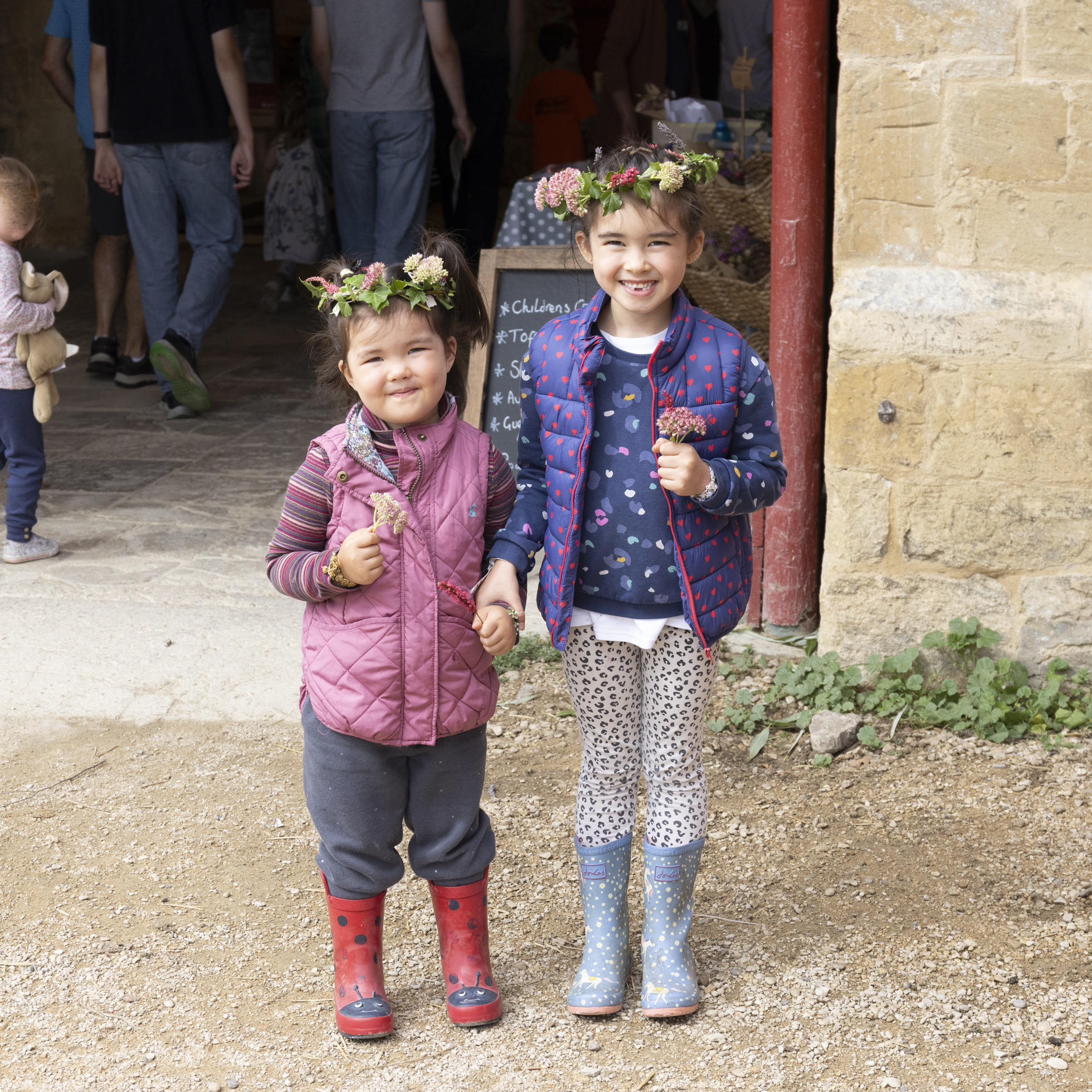 Two young children in flower crowns and wellies, holding posies smiling 